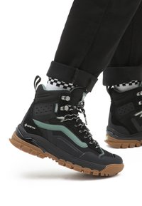 Czarno-zielone wysokie buty górskie z teksturowaną cholewką, grubą gumową podeszwą i wzorzystymi sznurówkami; wyposażone w Gore-Tex dla wodoodporności.