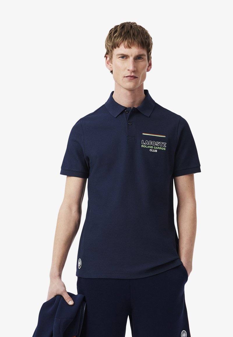 Mörkblå piké med ett texturerat tyg, treknapps-knäppning och broderad "LACOSTE ROLAND GARROS CLUB" logotyp på bröstet.