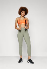 Fényes narancssárga sportmelltartó fehér márkázással, olajzöld, magas derekú leggings-el párosítva. A modell ugrókötéllel a kezében áll, fekete sportcipőt visel.