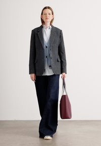 Blazer a cuadros grises sobre un chaleco azul claro, camisa blanca y pantalones anchos oscuros. Sosteniendo un bolso de mano burdeos. Texturas de tela suaves.