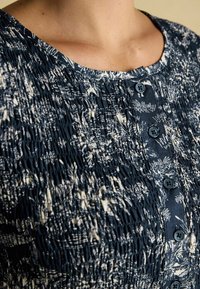 Námornícky modré šaty s textúrovaným smockingom a krémovým kvetinovým vzorom. Sú vybavené okrúhlym výstrihom a tmavými gombíkmi pozdĺž prednej časti.