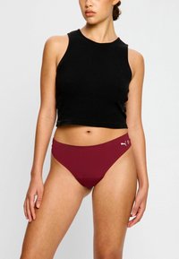 Bas de bikini bourgogne avec une taille haute, texture lisse et un petit logo blanc sur le côté. Le modèle porte un débardeur noir.