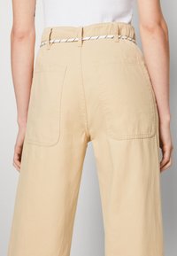 Béžové kalhoty s širokými nohavicemi a nařaseným pasem, doplněné kontrastním pásem černé a bílé barvy a dvěma zadními kapsami s kulatými rohy.