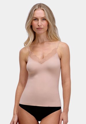 Blonde vrouw die een nudekleurige top met kant en zwart ondergoed draagt, staat tegen een effen lichte achtergrond en kijkt naar voren.