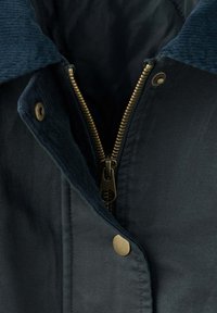 Veste sombre avec fermeture éclair en laiton partiellement ouverte et boutons-pression, avec une doublure de col bleu foncé texturée.