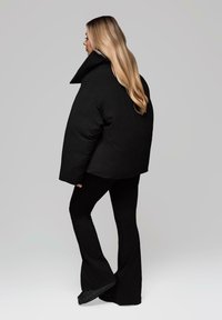 Černá péřová bunda s vysokým límcem, over-sized střihem a hladkou texturou, v kombinaci s rozšířenými černými kalhotami a plochými černými botami.