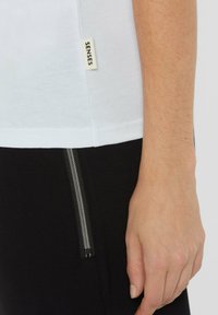 Weißes Baumwoll-T-Shirt mit einem kleinen Markenlabel an der Seite; kombiniert mit schwarzen Jogginghosen, die einen silbernen Reißverschluss zeigen.