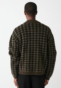 Brūns un melns rūtainais adīts kardigāns, kas ir ar apkakli, ribotu apakšmalu un teksturētu audumu. Brīva piegriezuma ar nolaižamām plecu šuvēm.