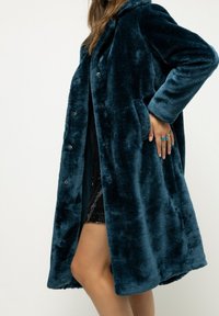 Čalouněný kabát z imitace kožešiny v modrozelené barvě s kapucí, volným střihem, bočními kapsami a zapínáním na patentky. Měkká textura zvyšuje teplo a pohodlí.
