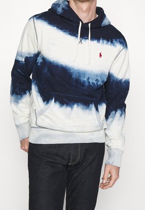Mann trägt einen marineblauen und weißen Tie-Dye-Pullover mit Kapuze und einem roten, gestickten Logo, kombiniert mit dunklen Jeans.