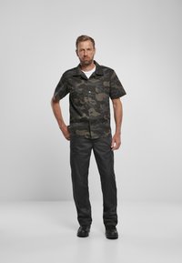 Camicetta a maniche corte con stampa camouflage verde scuro e nera, dotata di due taschini sul petto. Abbinata a pantaloni neri e scarpe nere.