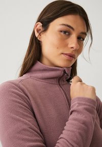 Fleecejakke i blød mauve med høj krave, halv lynlås og diskret branding på ærmet. Har ribbede manchetter og tekstureret stof.