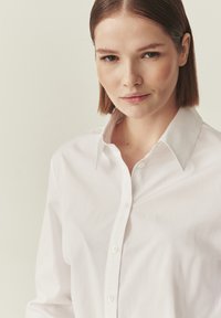 Chemise blanche à boutons avec un col classique, confectionnée dans un tissu léger. Texture lisse avec un léger éclat, dotée d'une patte de boutonnage soignée.