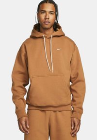 Felpa in cotone marrone con tasca frontale, cordini bianchi e piccolo logo Nike bianco sul petto. Vestibilità rilassata con polsini a coste.