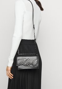 Borsa a tracolla nera in pelle con catena argentata, caratterizzata dal nome "Karl" in rilievo corsivo sulla parte anteriore. Texture liscia, forma rettangolare.