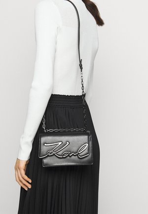 Bolso de cuero negro cruzado con cadena de plata, que presenta el nombre "Karl" en cursiva en relieve en la parte frontal. Textura suave, forma rectangular.