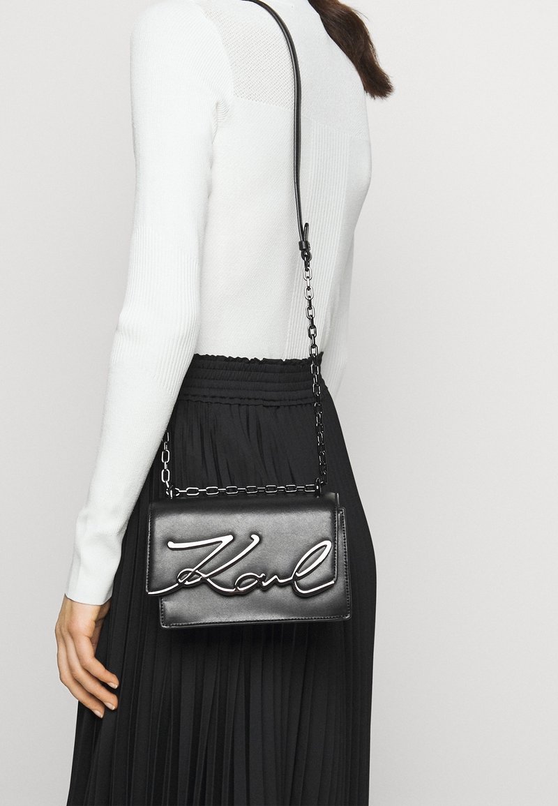 Borsa a tracolla nera in pelle con catena argentata, caratterizzata dal nome "Karl" in rilievo corsivo sulla parte anteriore. Texture liscia, forma rettangolare.
