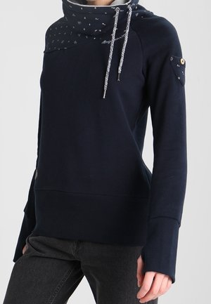 Vrouw draagt een donkerblauwe sweatshirt met een hoge, patroonrijke kraag en trekkoorden, lange mouwen en een afgeknoopte zak op de bovenarm.