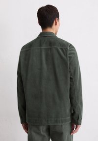 Zielona kurtka z aksamitu prążkowanego o strukturalnym kroju, z widocznym prążkowaniem, kołnierzem i mankietami zapinanymi na guziki, przedstawiona od tyłu.