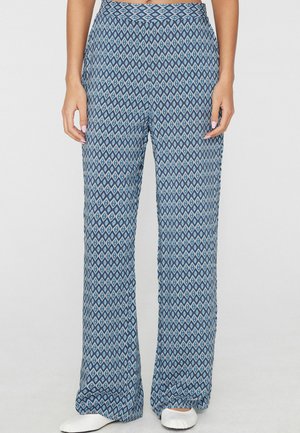 Frau trägt hoch taillierte, weit geschnittene Hose mit blauem geometrischem Diamantmuster und weißen flachen Schuhen, steht vor einem einfarbigen Hintergrund.