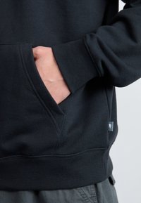 Haut à capuche noir avec une poche kangourou, des manches et un ourlet côtelés. Tissu texturé avec des détails cousus et une petite étiquette de logo sur le côté.