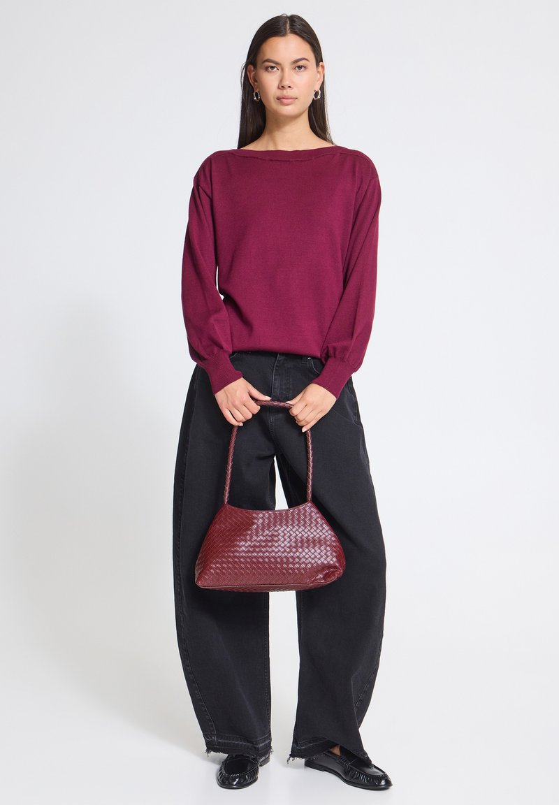 Femme portant un pull bordeaux et un jean noir à jambes larges, tenant un sac à main tissé bordeaux assorti devant un fond blanc uni.