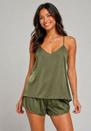Femme souriante portant un caraco en satin vert olive et un short assorti, debout devant un fond gris uni.