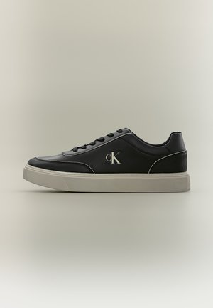 Scarpa da ginnastica bassa nera con suola bianca, lacci neri e logo CK argento sul lato, mostrata di profilo su sfondo semplice.