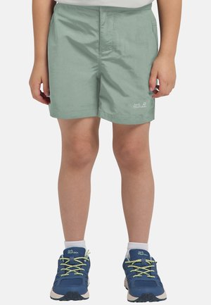 Kind trägt salbeigrüne Jack Wolfskin Shorts, weiße Socken und blaue Wanderschuhe mit gelb-grünen Schnürsenkeln, steht vor weißem Hintergrund.