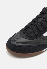 Svarta sneakers med en överdel i läder och mocka, med en texturerad svart sula och vaxade svarta snören. Vita detaljer på sidan.