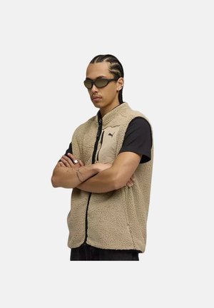 Uomo con capelli intrecciati e occhiali da sole che indossa un gilet in pile beige sopra una camicia nera, in piedi con le braccia incrociate su uno sfondo uniforme.