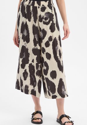 Vrouw met wijde, cropped broek met een zwarte en beige abstracte luipaardprint en zwarte open-sandalen met gouden gespen.