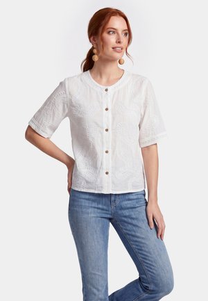 Femme rousse portant un chemisier blanc brodé à manches courtes et boutonné ainsi qu'un jean bleu, debout avec une main sur la hanche devant un fond clair.