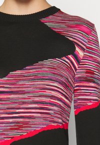 Gros plan d'un pull en tricot multicolore avec des rayures horizontales noires, roses, violettes, rouges et blanches, et un motif triangulaire noir audacieux près de l'épaule.