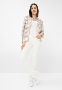 Beigestreiftes Hemd mit weißen Streifen, mit Knopfverschluss; kombiniert mit weißen, ausgestellten Jeans und weißen Sneakern. Das Model posiert mit einer Hand in der Tasche.