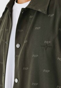 Giacca camicia verde oliva realizzata in tessuto liscio, con bottoni a scatto neri, un sottile motivo "PXP" e un dettaglio di stella ricamata.