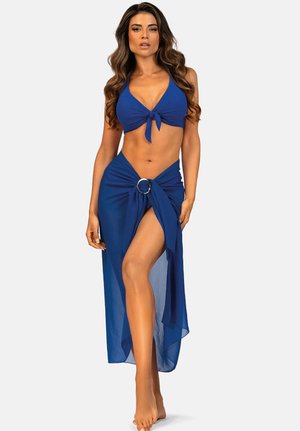 Kobieta o długich falujących włosach, ubrana w niebieski top bikini z wiązaniem z przodu oraz pasującą przezroczystą spódnicę sarong z ozdobnym pierścieniem, pozująca na boso.