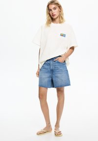 Krémové oversized tričko s barevným potiskem, spárované s světle modrými džínovými šortkami s třepenými lemy. Model nosí žabky.