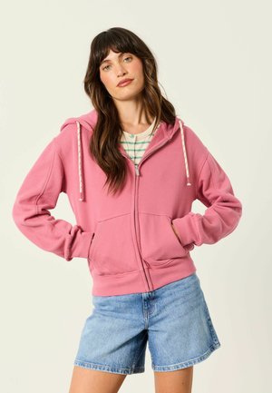 Frau mit langen braunen Haaren trägt einen rosa Kapuzenpullover mit Reißverschluss über einem gestreiften Shirt und Jeansshorts, steht mit den Händen in den Taschen.