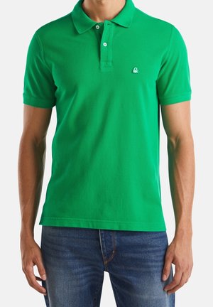 Muž, který má na sobě jasně zelené polo tričko s krátkým rukávem se dvěma knoflíky, malé logo na levé hrudi, spárované s modrými džínami.