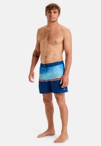 Mężczyzna stojący boso, ubrany w niebieskie, gradientowe spodenki kąpielowe ze sznurkiem w talii i z markowym paskiem, na jednolitym jasnym tle.