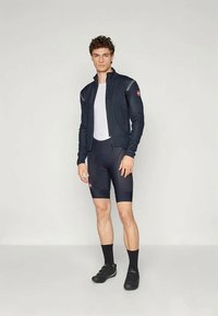 Completo da ciclismo maschile: giacca nera aderente a maniche lunghe, sottogiacca bianca, pantaloni corti neri con dettagli a pannello e scarpe da ciclismo nere.