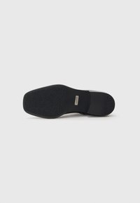 Suela de zapato de cuero negro con una parte inferior de goma texturizada, que presenta un ligero tacón y una placa metálica de logo cerca del centro.