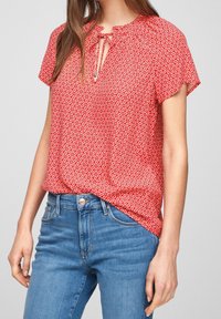 Blouse rouge à manches courtes avec un motif floral, encolure goutte d'eau, détail froncé et coupe décontractée, associée à un jean bleu taille haute.