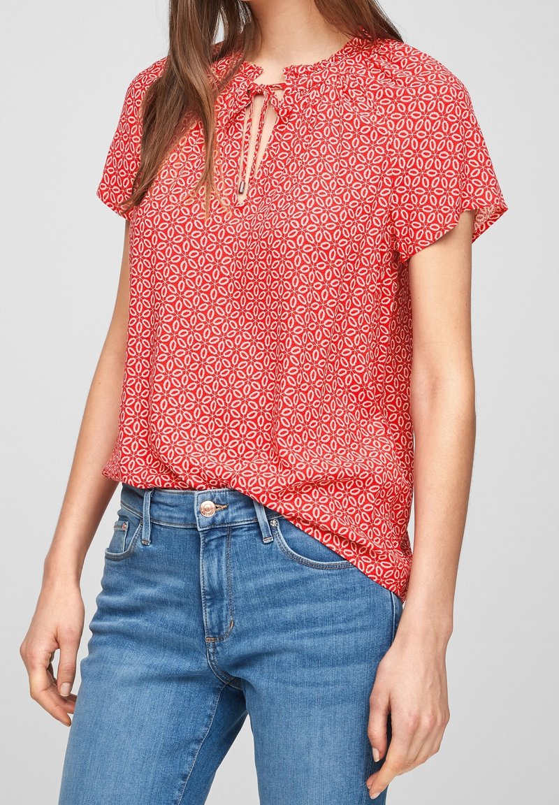 Blouse rouge à manches courtes avec un motif floral, encolure goutte d'eau, détail froncé et coupe décontractée, associée à un jean bleu taille haute.