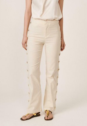 Mujer con pantalones de tiro alto color crema con botones dorados a lo largo de las costuras laterales, combinados con una camiseta sin mangas blanca y sandalias negras con detalles dorados.