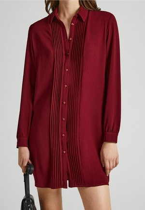 Femme portant une robe boutonnée bordeaux à manches longues avec des plis verticaux sur le devant, tenant un sac à main noir dans une main.