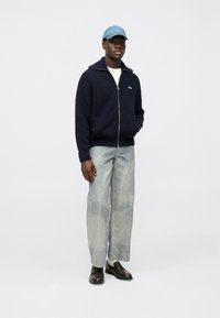 Marineblå genser med glidelås, lyseblå cap, hvit skjorte, lystvaskede teksturerte jeans og mørkebrune loafers. Minimalistisk design.