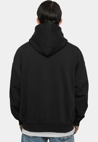 Black hooded sweatshirt with a relaxed fit, ribbed cuffs and hem, and soft texture. Worn over a white t-shirt, seen from the back.