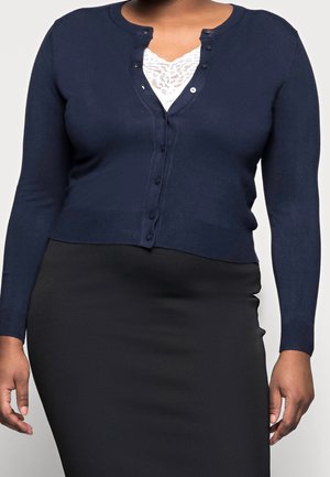 Cardigan marine à manches longues, à col rond et avec des fermetures à boutons. Confectionné en tissu tricoté lisse, associé à un haut en dentelle blanche en dessous.